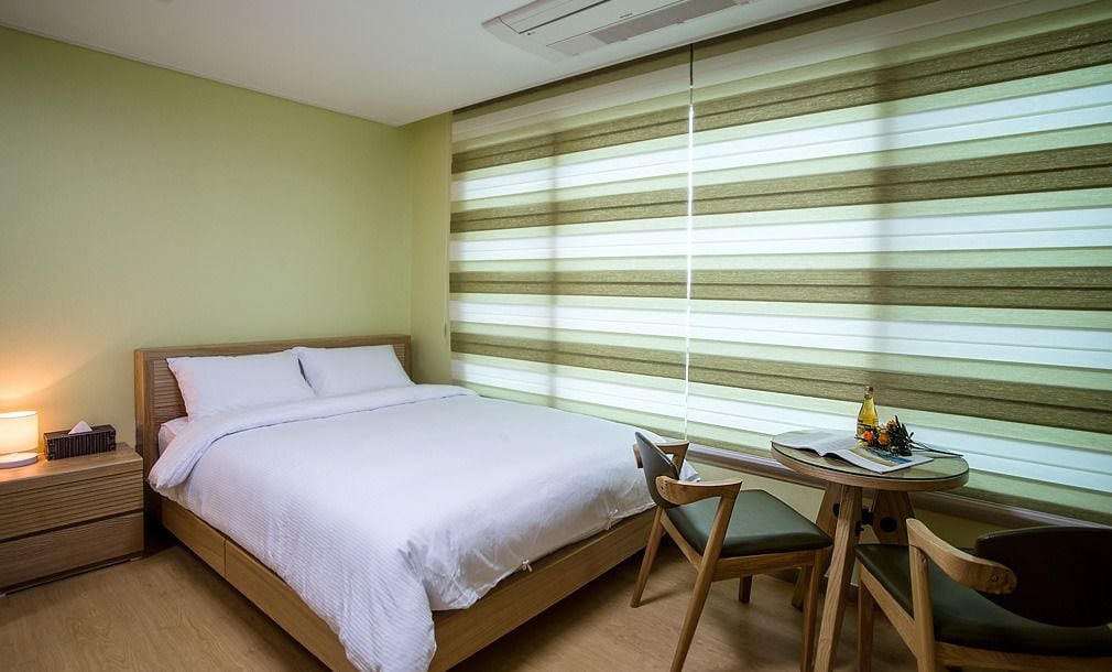 Quarto de hotel com cama, mesa de cabeceira, abajur e uma pequena mesa redonda com cadeiras.