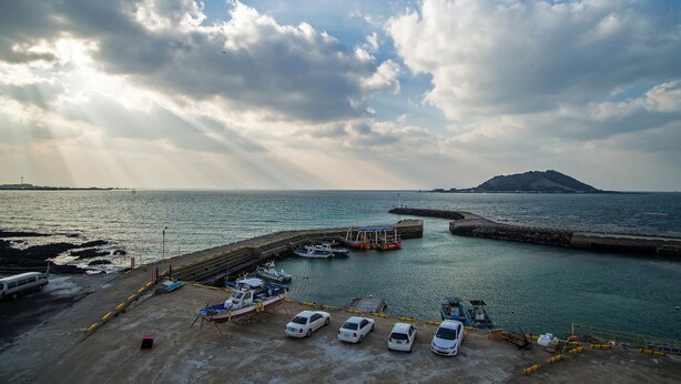 A coastal marina with parked cars and boats, a pier extending into the water, and a distant island. - Sleep in the Sea (Jeju City)