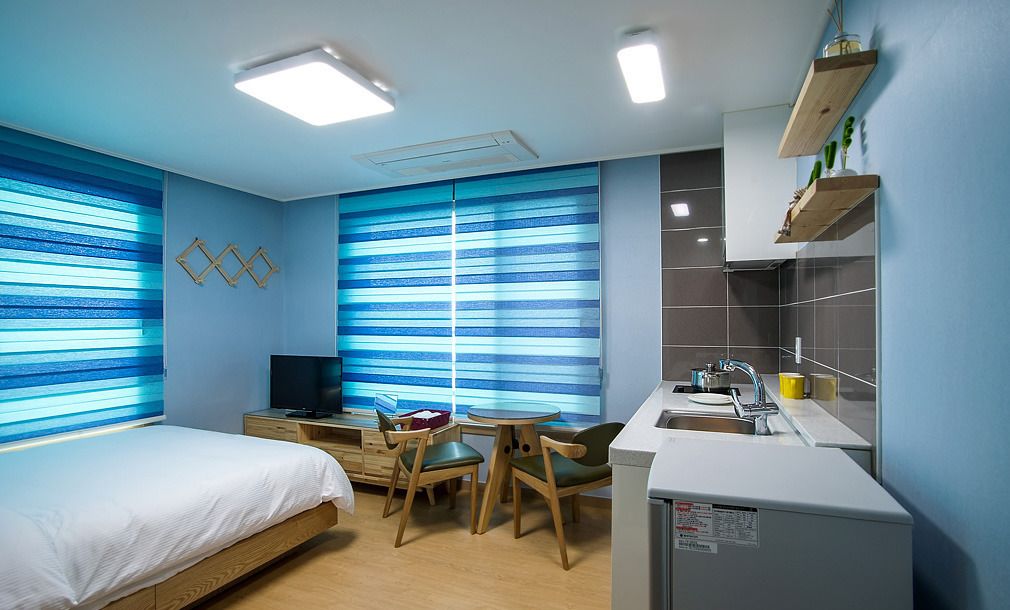 A compact hotel room with a bed, a desk with a chair, a TV, a small kitchen area with a sink and a refrigerator, and large windows with blue-striped blinds.