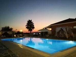 A swimming pool with a stone border, surrounded by lounge chairs and umbrellas, with a sunset in the background. - Saint George Hotel & Rooms (Karystos)