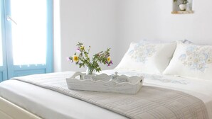A bed with white bedding and floral pillows, a white decorative tray with flowers, and a small plant on a bedside table. - Nakos Homes (Paros)