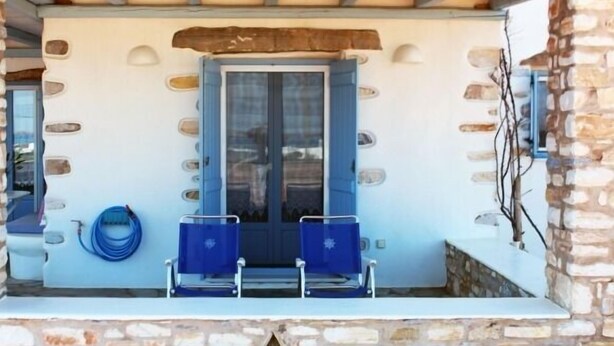 A stone-walled porch with a blue door, two blue chairs, and a hose on the wall. - Nakos Homes (Paros)