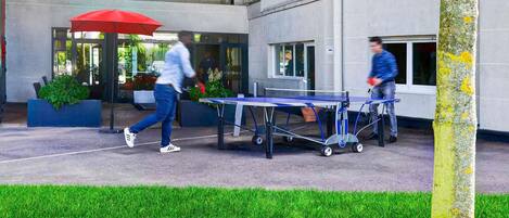 An outdoor area with a ping pong table, a red umbrella, and a building entrance.