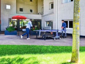 An outdoor area with a ping pong table, a red umbrella, and a building entrance.