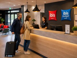 Una recepción de hotel con la marca 'ibis Hotels', una recepcionista e invitados.