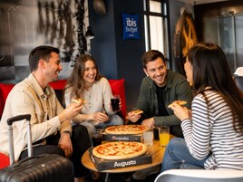 A group of people sitting around a table with pizzas and drinks, in a room with a blue wall and a sign that reads 'ibis budget.'