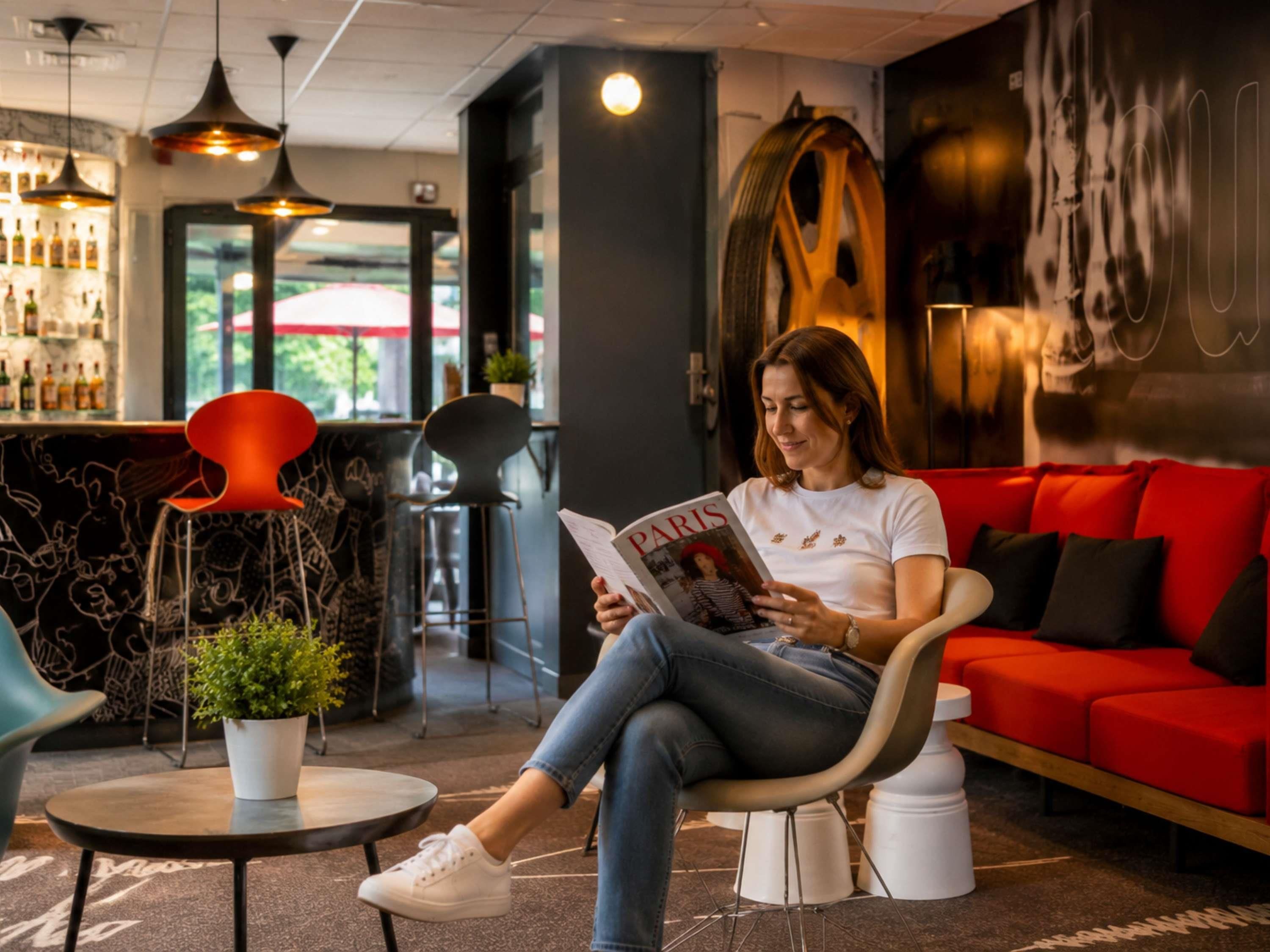 Una mujer leyendo una revista en un área de descanso moderna con un sofá rojo, una barra con botellas y una gran obra de arte abstracta en la pared.