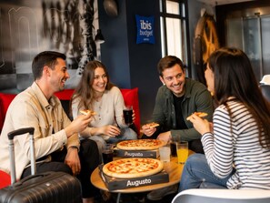 Un grupo de personas sentadas alrededor de una mesa con pizzas y bebidas, en una habitación con pared azul y un cartel que dice 'ibis budget'.