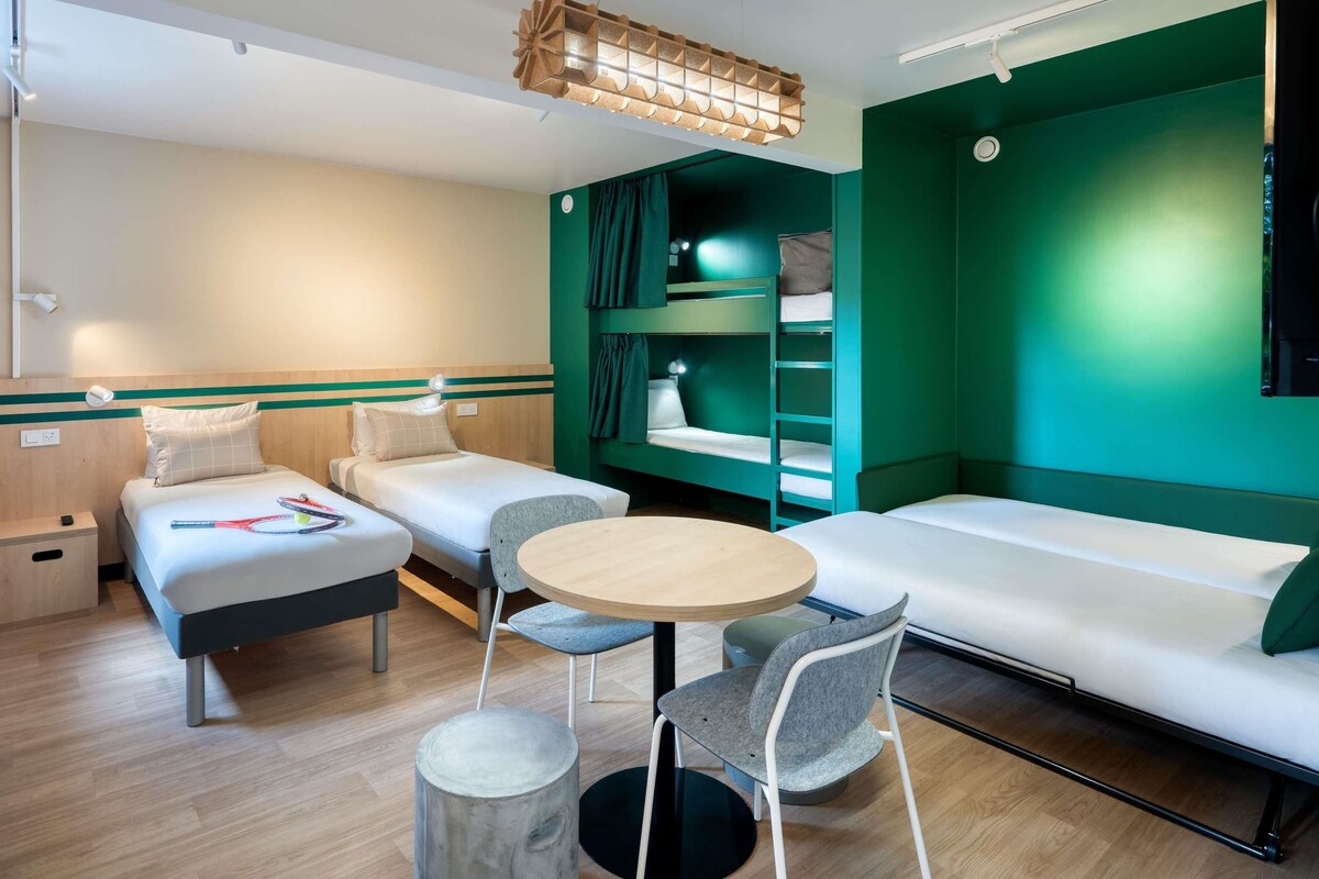 A compact hotel room with two single beds, a sofa bed, a small round table, and a chair. There is a bunk bed with a ladder and a hanging light fixture above the beds.