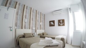 A hotel room with two beds, a wooden wall panel, an air conditioning unit, and a window with curtains. - Hostal del Mar (Tossa de Mar)