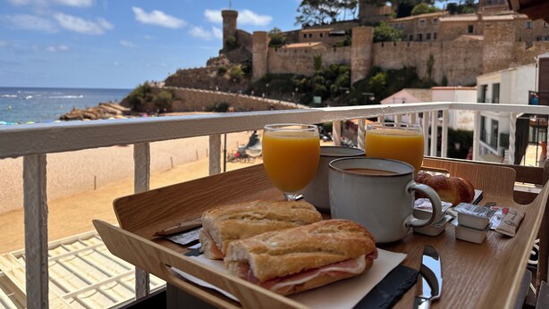 A breakfast tray with sandwiches, juice, and coffee on a balcony overlooking a coastal town with historic buildings. - Hostal del Mar (Tossa de Mar)