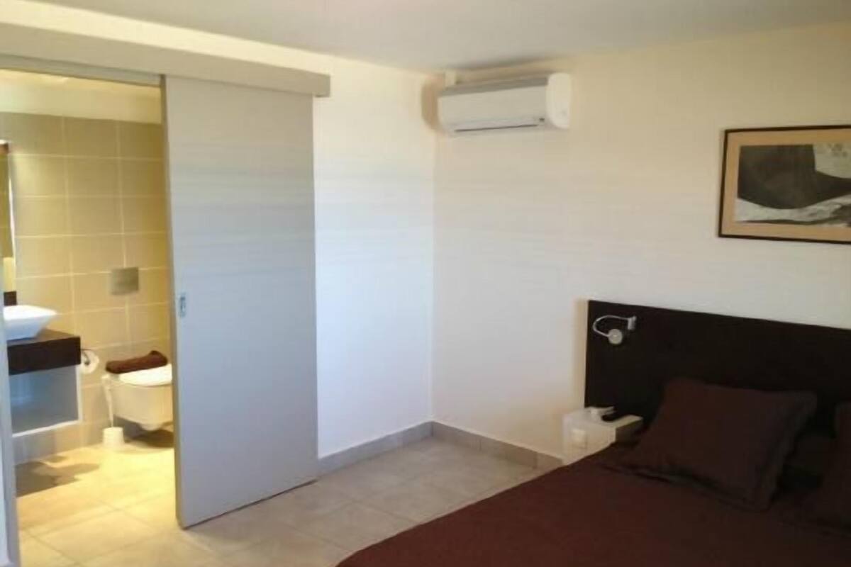 Quarto de hotel com cama, mesa de cabeceira, ar condicionado fixado na parede e banheiro visível por uma porta aberta.