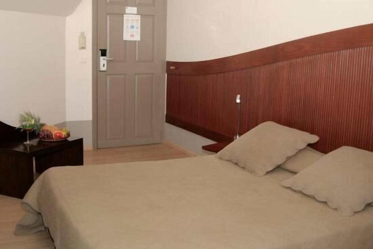 Quarto de hotel com cama, mesa de cabeceira, porta e uma pequena mesa com frutas e uma planta.
