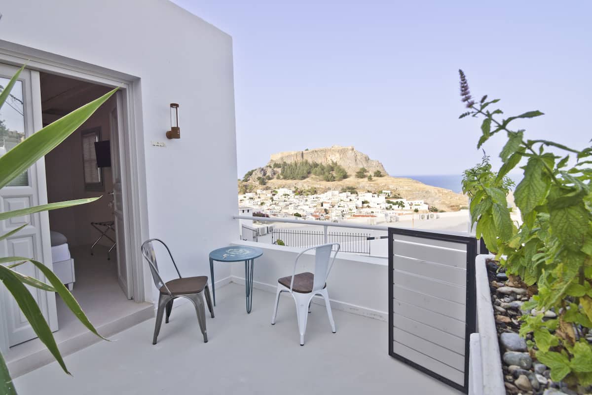 A balcony with two chairs and a small table, overlooking a coastal town and a hill with a fort.