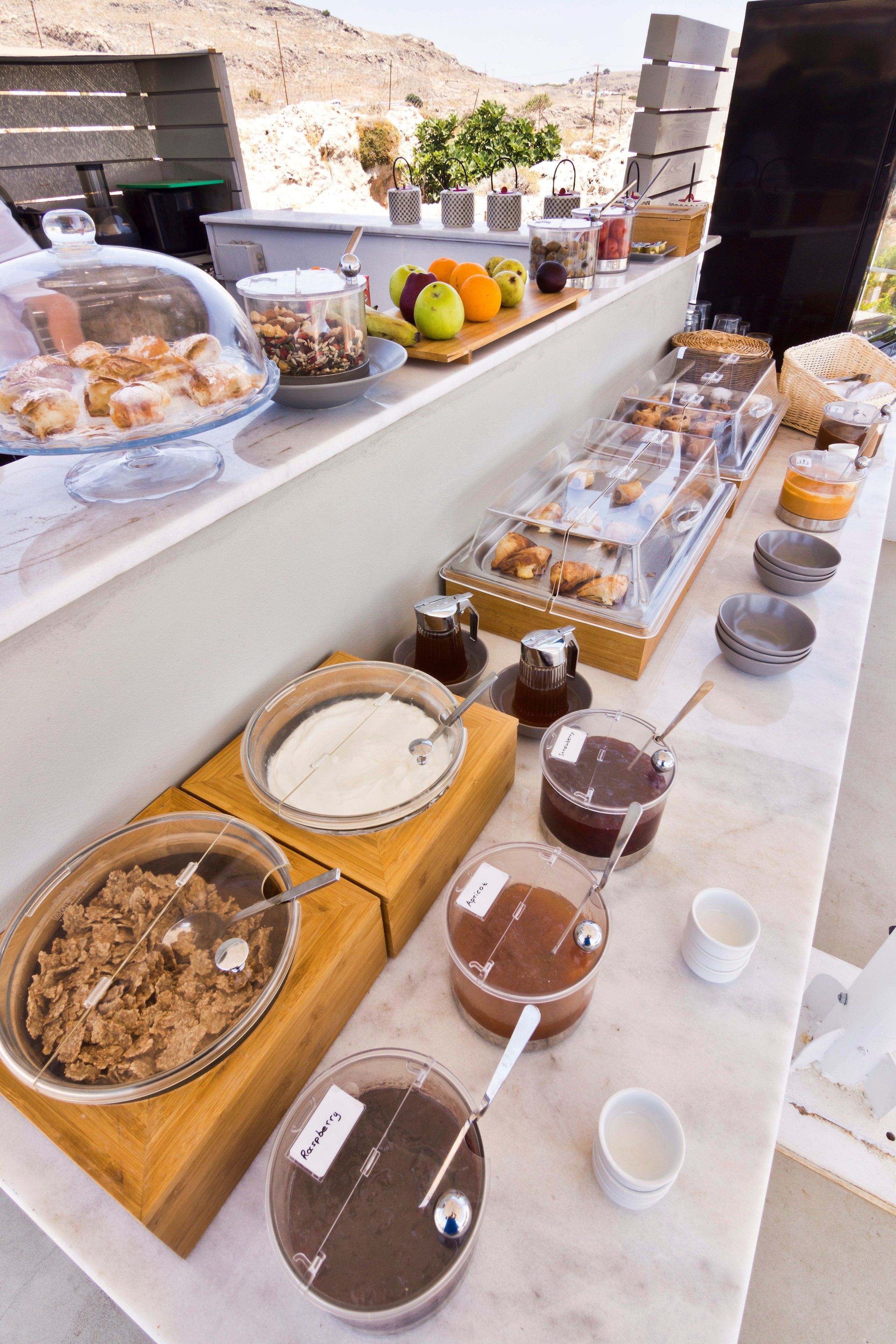 A buffet with various food items and beverages, including fruits, pastries, and condiments, arranged on a marble counter.