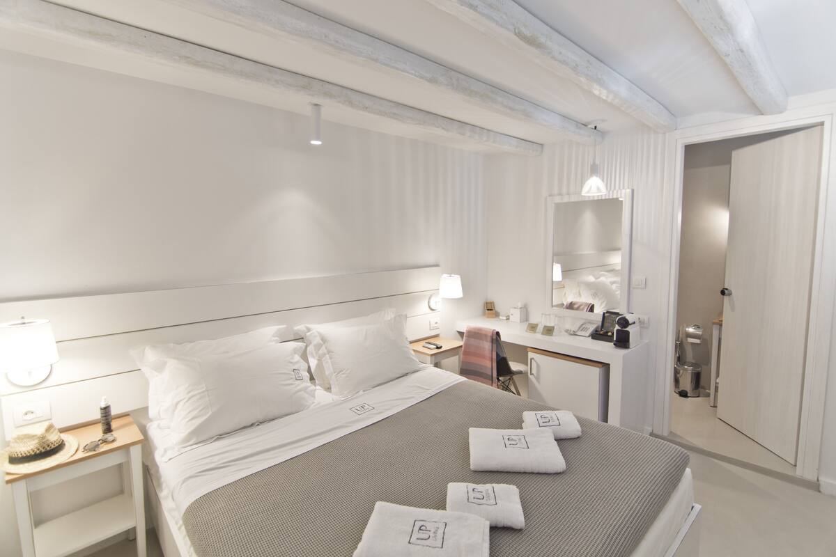 A hotel room with a neatly made bed, a bedside table with a lamp, a desk with a chair, and a mirror above the desk.