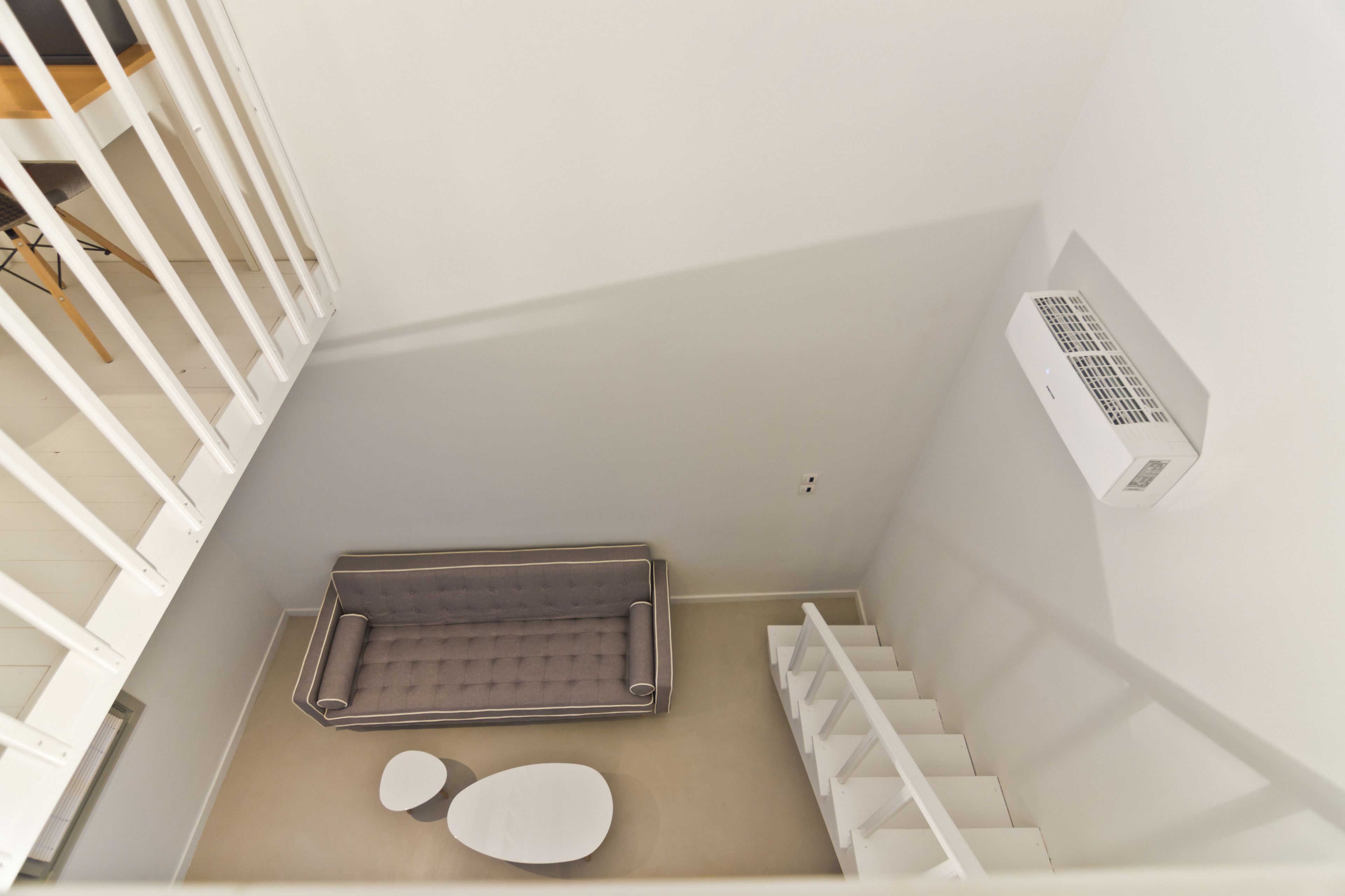 Uma escadaria moderna e minimalista com corrimão branco, uma pequena área de estar com um banco acolchoado e uma mesa redonda, e um ar condicionado fixado na parede.