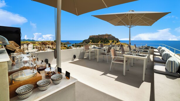 A rooftop terrace with outdoor seating, tables, and umbrellas, overlooking a coastal landscape with a hill and the sea. - Lindos Comfy Suites (Rhodes)