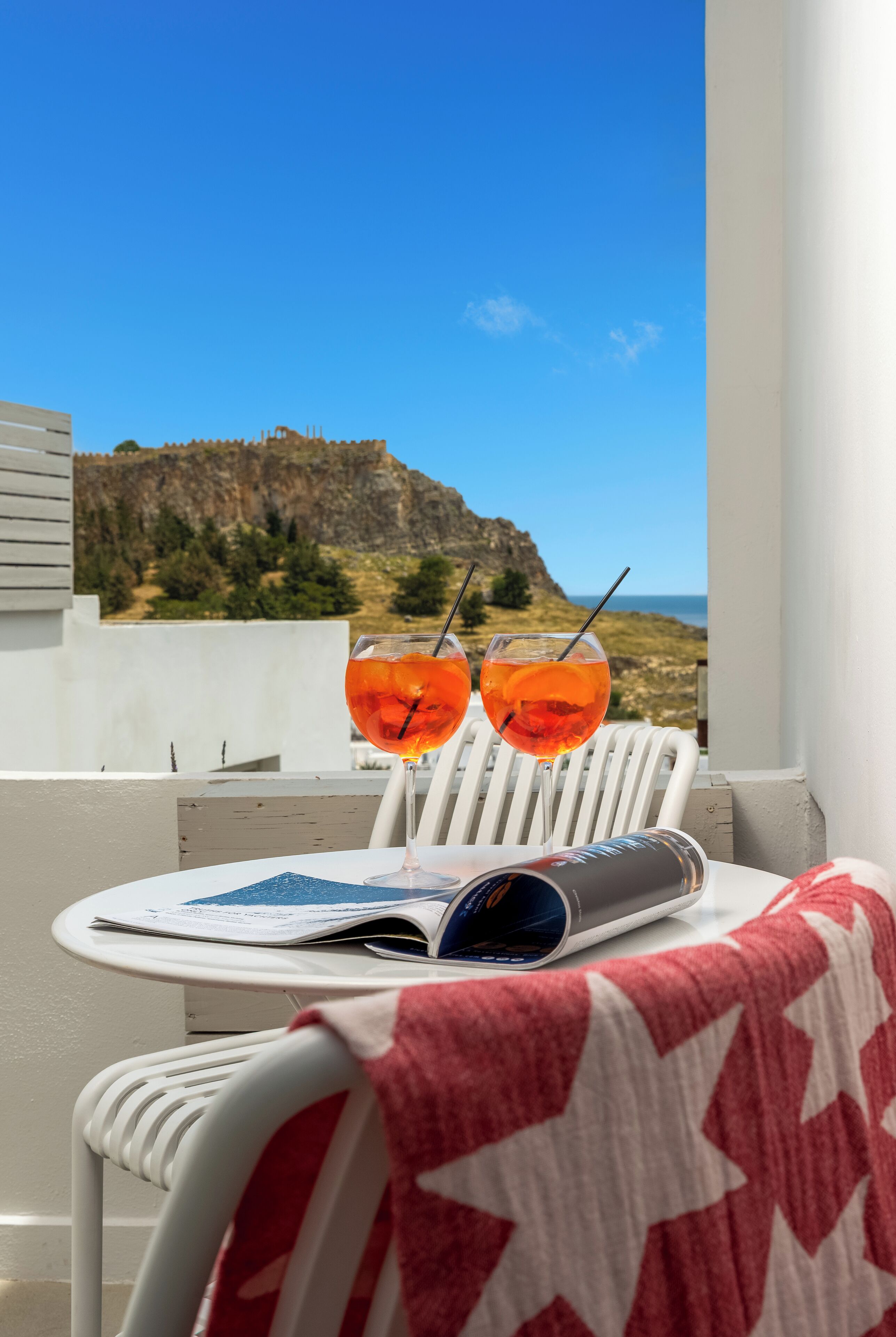 Un balcón con dos sillas blancas, una mesa con una revista y dos vasos de jugo de naranja.