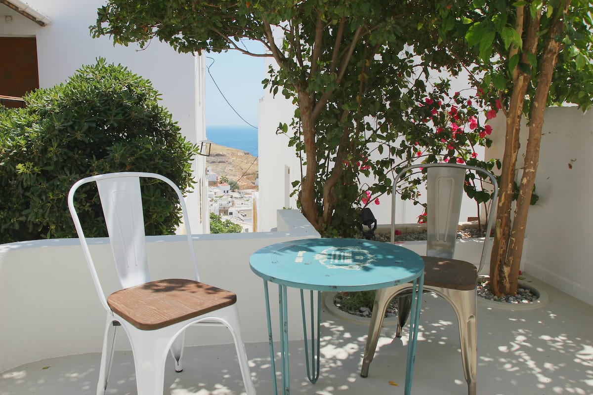 A balcony with a white chair and a turquoise table, overlooking a coastal area with white buildings and a tree with pink flowers.