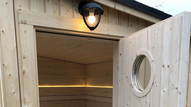 Un sauna avec un intérieur en bois, un banc et un luminaire au-dessus de l’entrée.