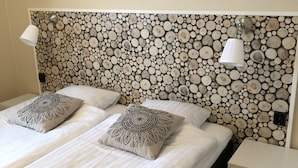 Un lit double agrémenté de deux coussins décoratifs, une table de chevet avec une lampe et un mur orné d’un motif circulaire en bois.