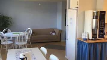 A dining area with a table set for two, a coffee maker, and a bowl of food. There is a sofa and a plant in the background.