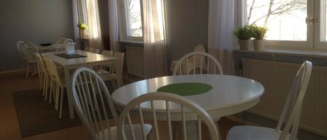 A dining area with white chairs and a round table, large windows with curtains, and a potted plant on the windowsill.