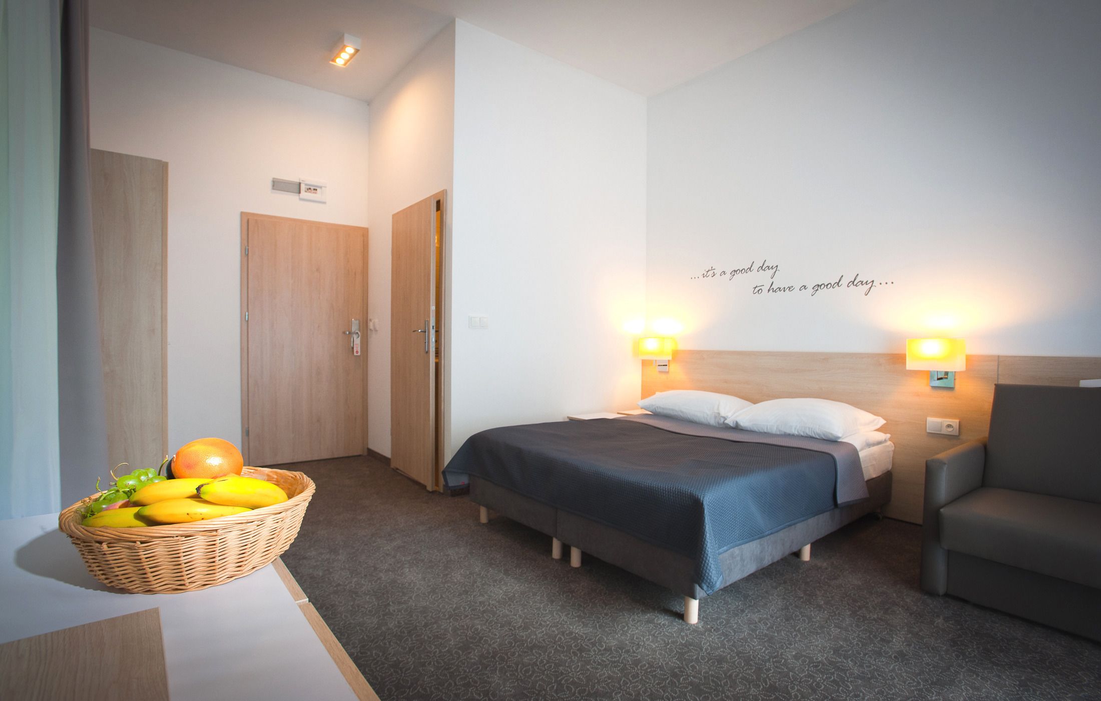 A hotel room with a bed, a chair, a wall with a motivational quote, a basket of fruit, and a door.