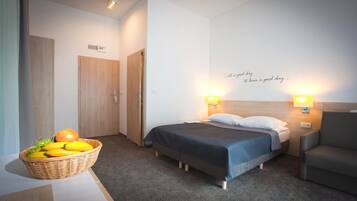 A hotel room with a bed, a chair, a wall with a motivational quote, a basket of fruit, and a door.