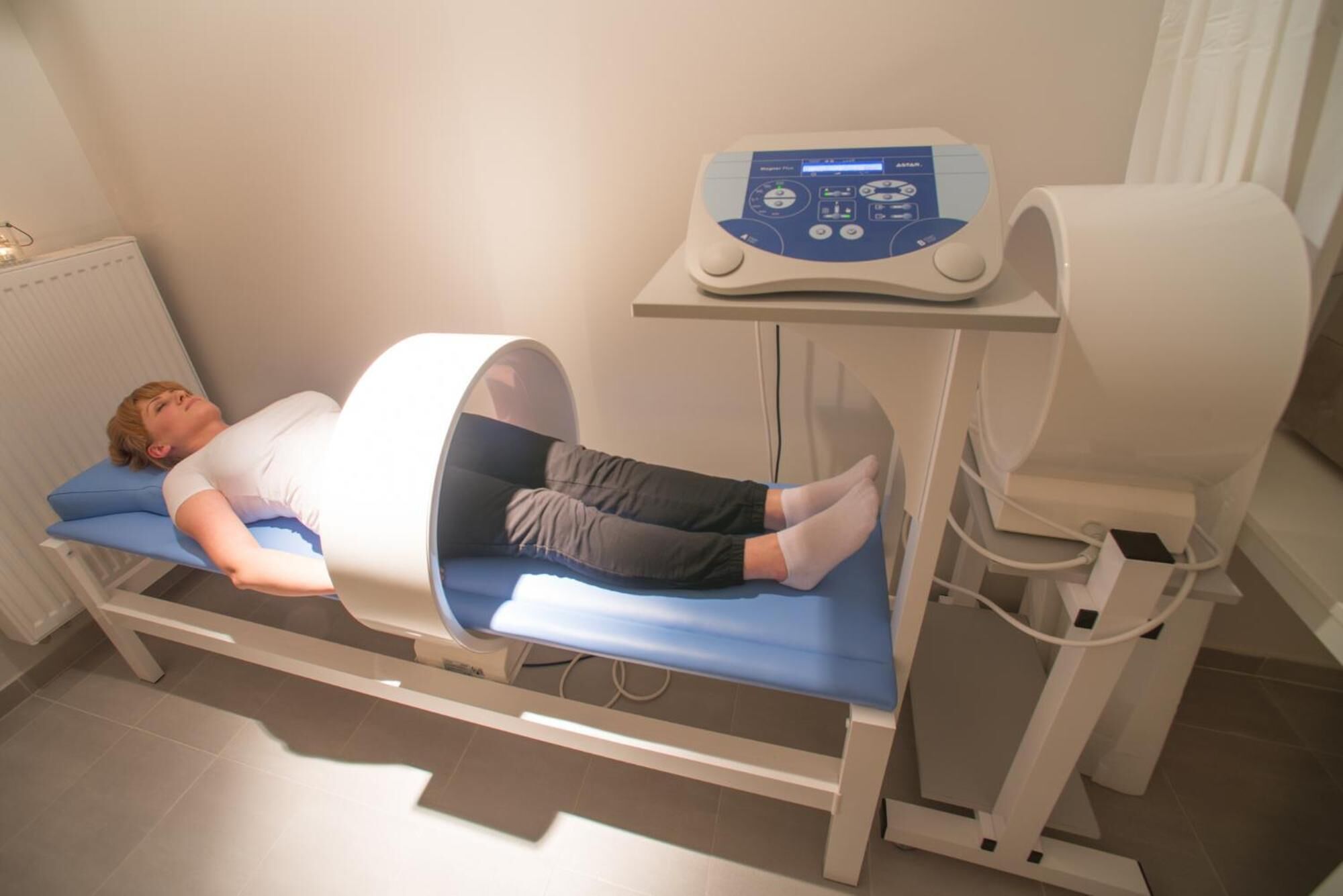 A medical imaging room with a patient lying on a bed, a control panel, and a chair.