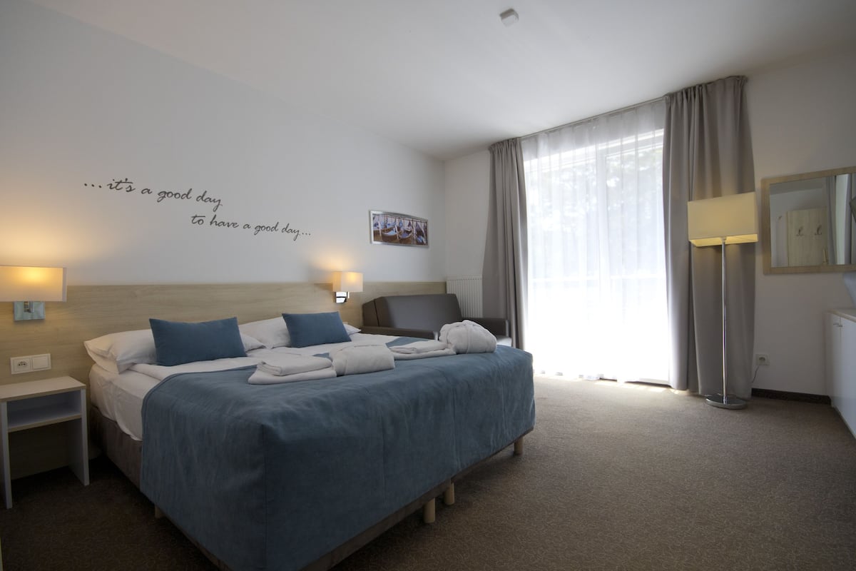 Una habitación de hotel con cama, mesitas de noche, lámpara, espejo y ventana con cortinas.