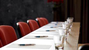Una mesa de conferencias con sillas rojas, adornada con bolígrafos y vasos, en una habitación con pared oscura y texturizada y un florero rojo.