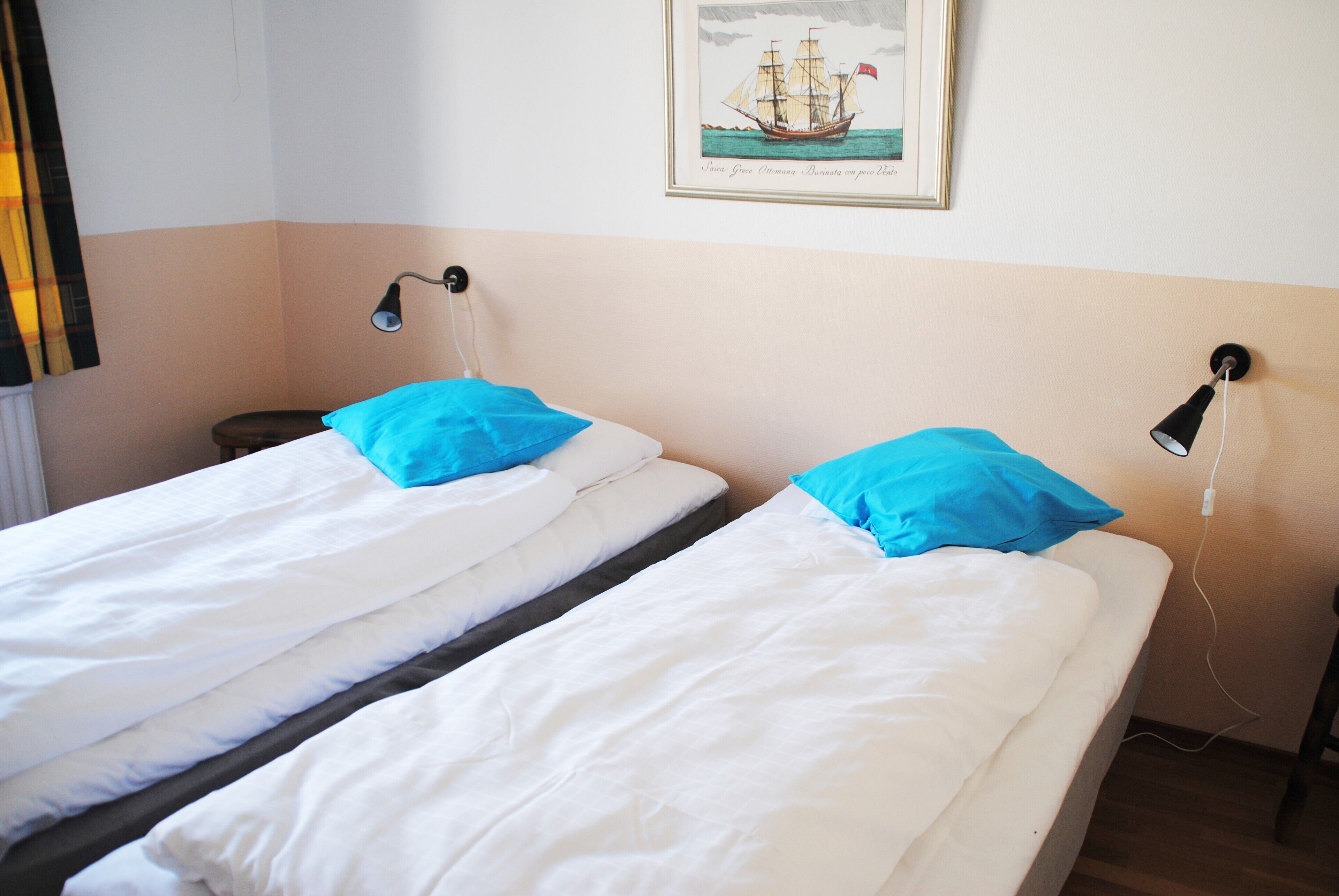 Two single beds with white bedding and blue pillows, a nightstand, a wall-mounted lamp, and a framed picture of a sailing ship.