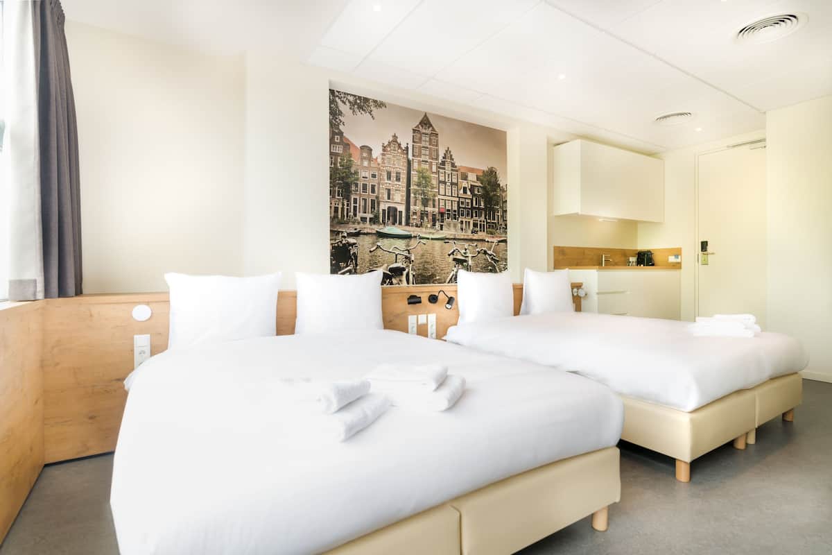 Ett hotellrum med två sängar, en sänggavel i trä, ett stort fönster med utsikt över byggnader och ett litet pentry.
