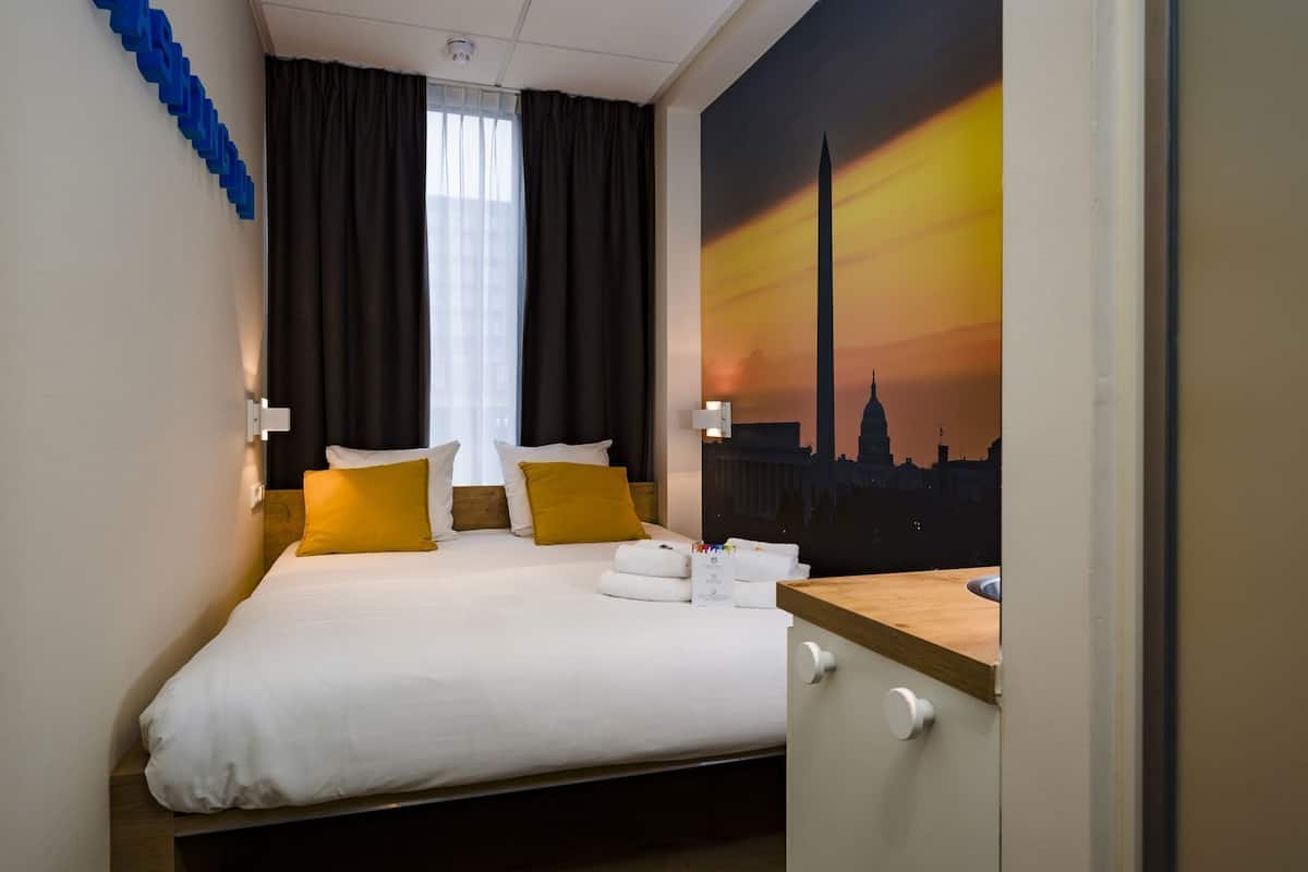 Ett hotellrum med en säng, två kuddar, ett nattduksbord, ett fönster med gardiner och en väggmålning.