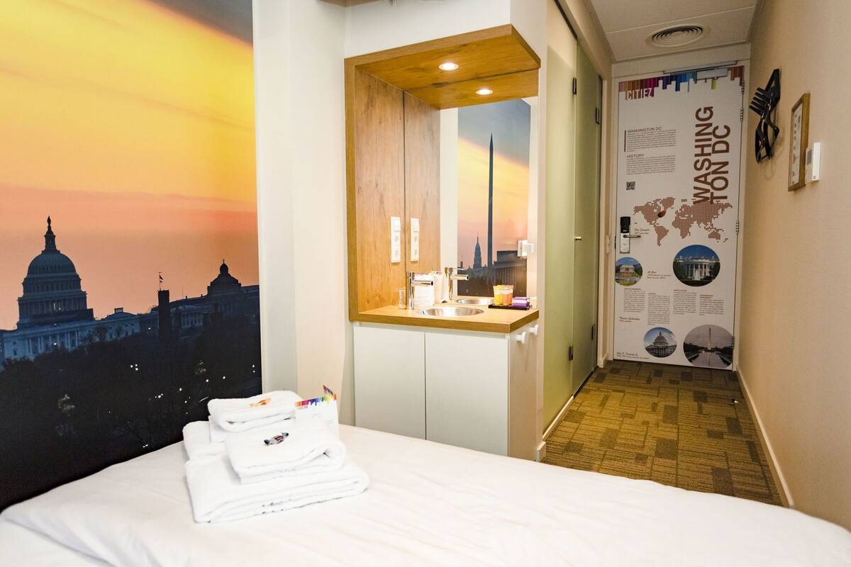 Ett hotellrum med en säng, ett badrum med en spegel, ett handfat och en vägg prydd med en affisch som föreställer en stadsbild.