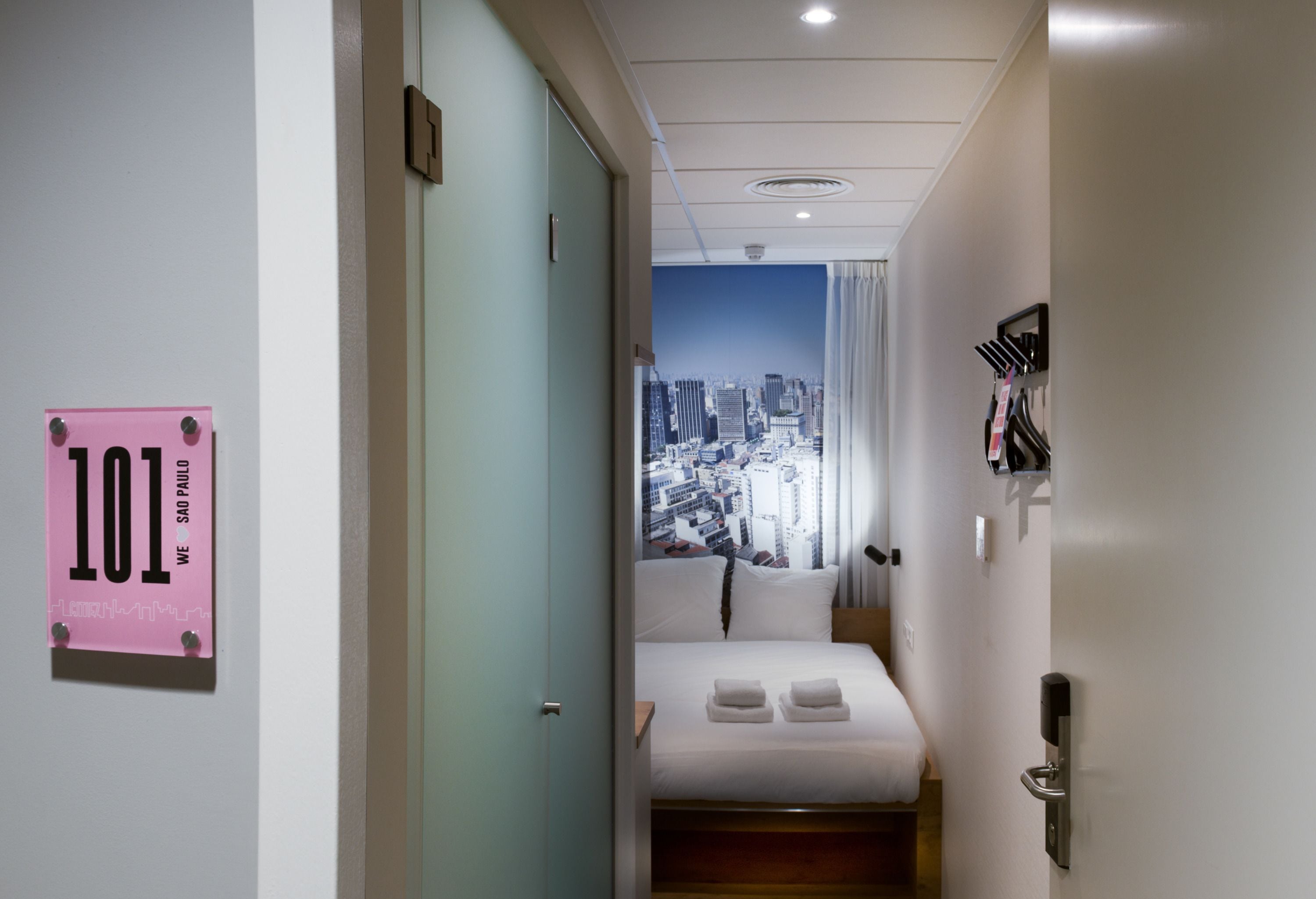 A hotel room with a cityscape view, a bed with white linens, a pink sign on the door, and a doorbell.
