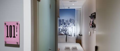 A hotel room with a cityscape view, a bed with white linens, a pink sign on the door, and a doorbell.