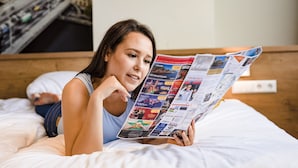 Una persona acostada en una cama con una revista, un almohadón y una pared con un cuadro de un paisaje urbano.