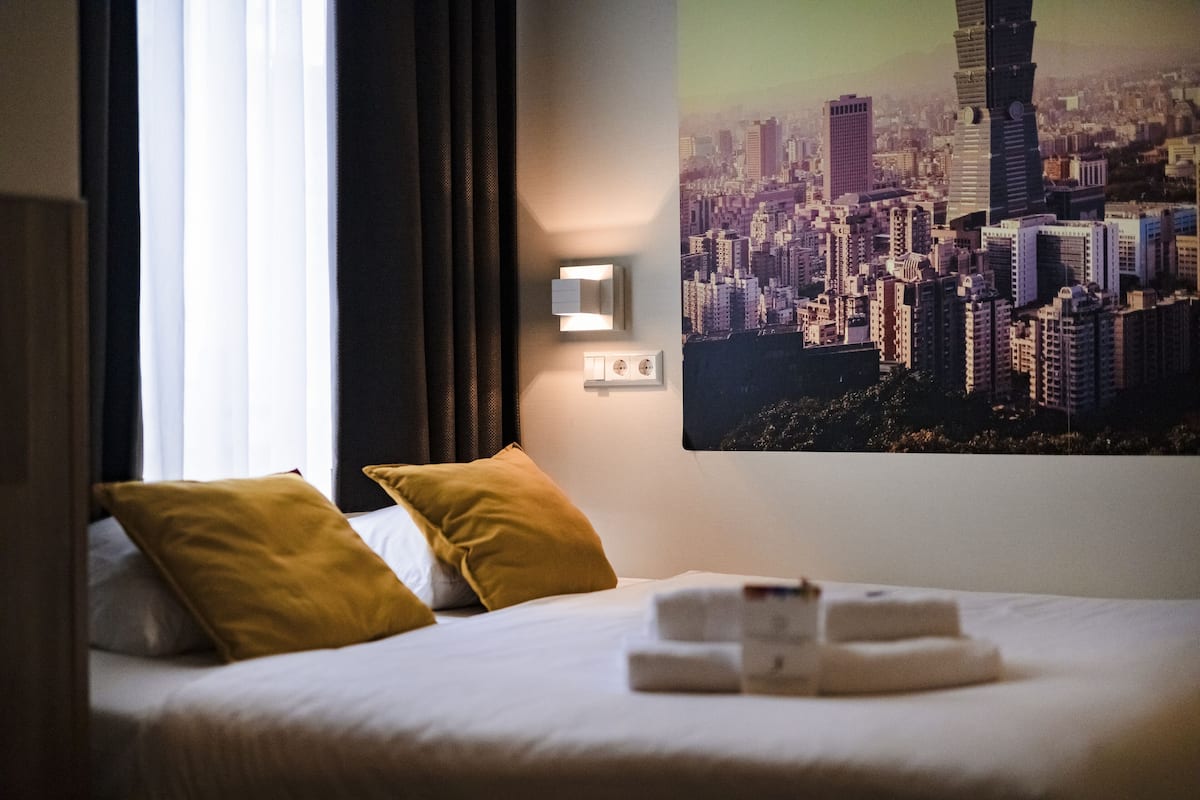 Ett hotellrum med en säng, två kuddar, en väggmonterad lampa och utsikt över staden.