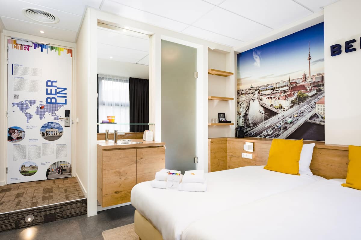 Ett hotellrum med en stor säng, en byrå i trä, en spegel och en vägg prydd med en affisch som föreställer en stadsbild.