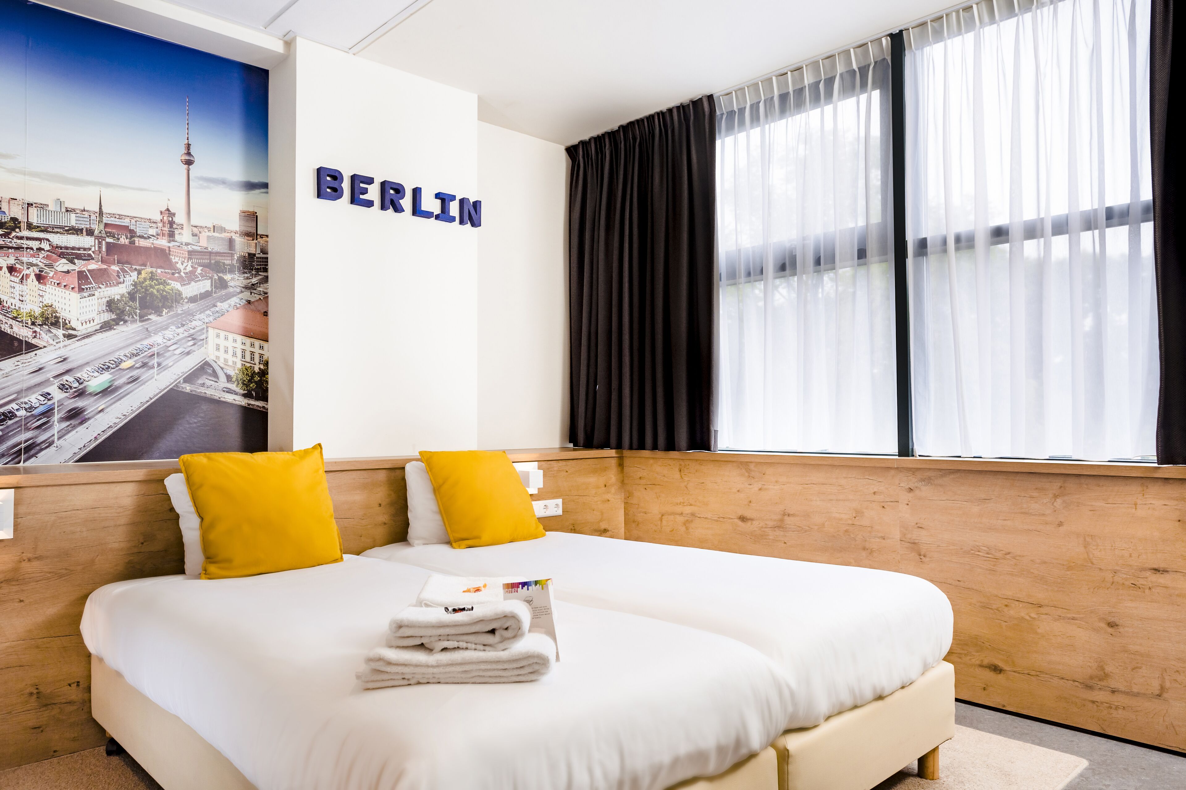 A hotel room with a large bed, two pillows, a neatly folded towel, a poster of Berlin, and a window with black curtains.