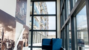 Un moderno vestíbulo de hotel con una escalera que conduce a un piso superior, grandes ventanas que ofrecen vistas de la ciudad y un sofá azul en el centro.