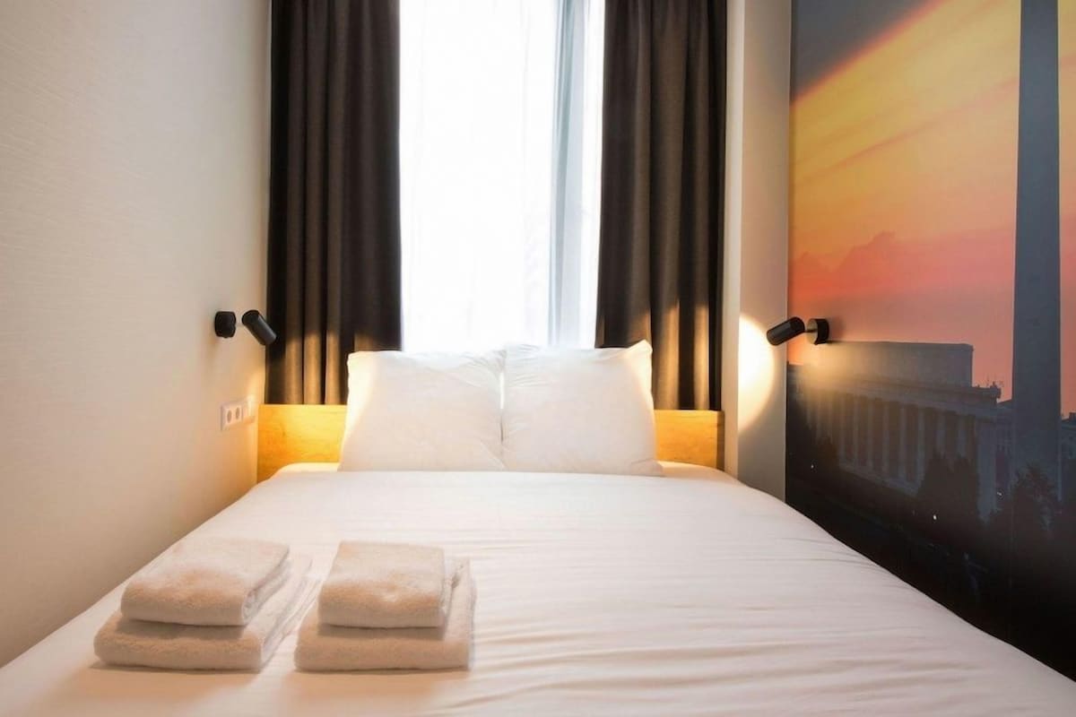 A hotel room with a bed, two pillows, a nightstand with a lamp, and a window with curtains.