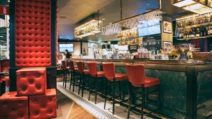 Un área de bar con taburetes tapizados en rojo, una barra y una variedad de botellas y vasos en estantes.