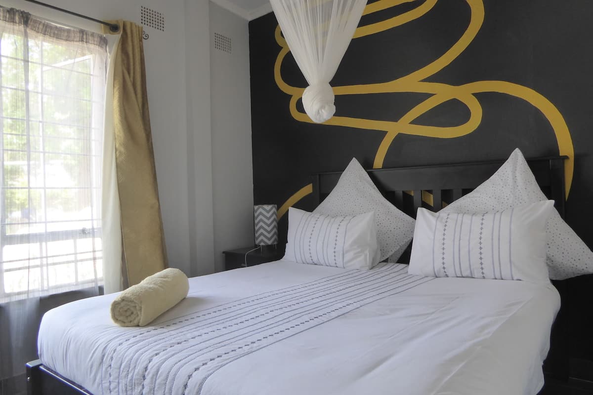 Un lit bien fait, avec du linge de lit blanc et une serviette enroulée, une tête de lit noire ornée d’un motif géométrique et une fenêtre avec des rideaux.