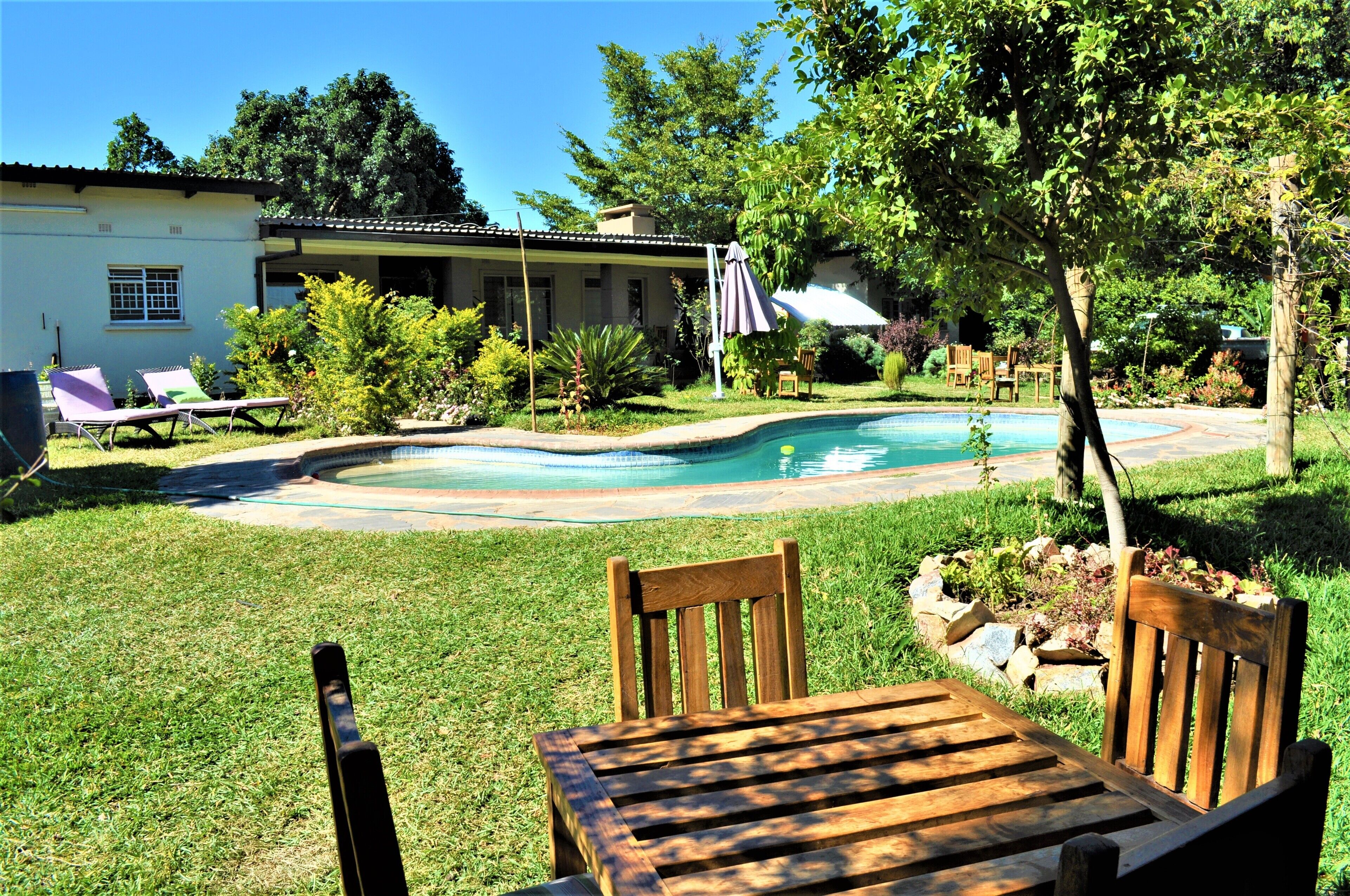 A backyard with a swimming pool, two lounge chairs, a wooden table, and a house in the background.