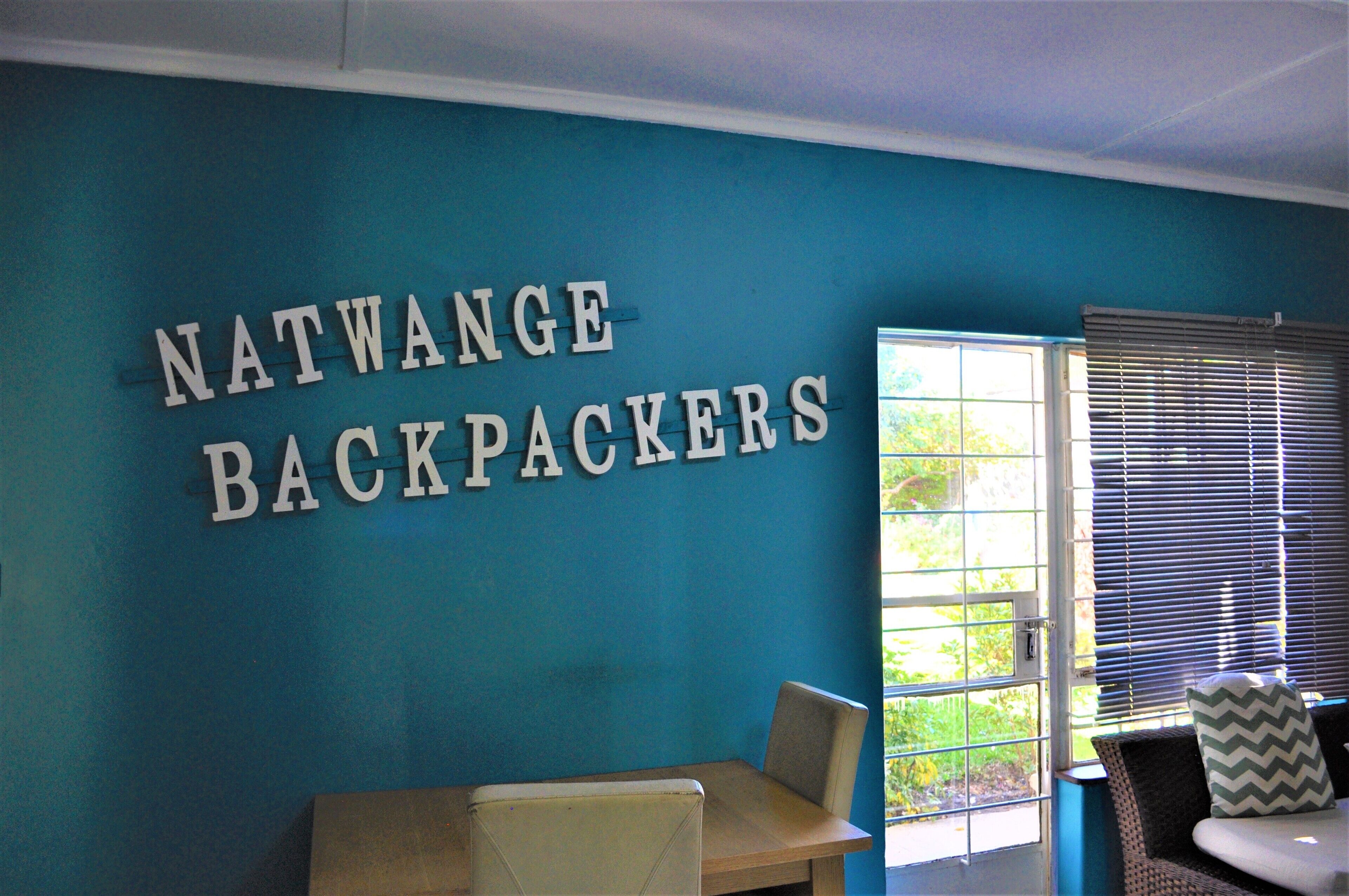 A room with a teal wall and a sign that reads "NATWANGE BACKPACKERS". There is a window with blinds, a chair, and a table.