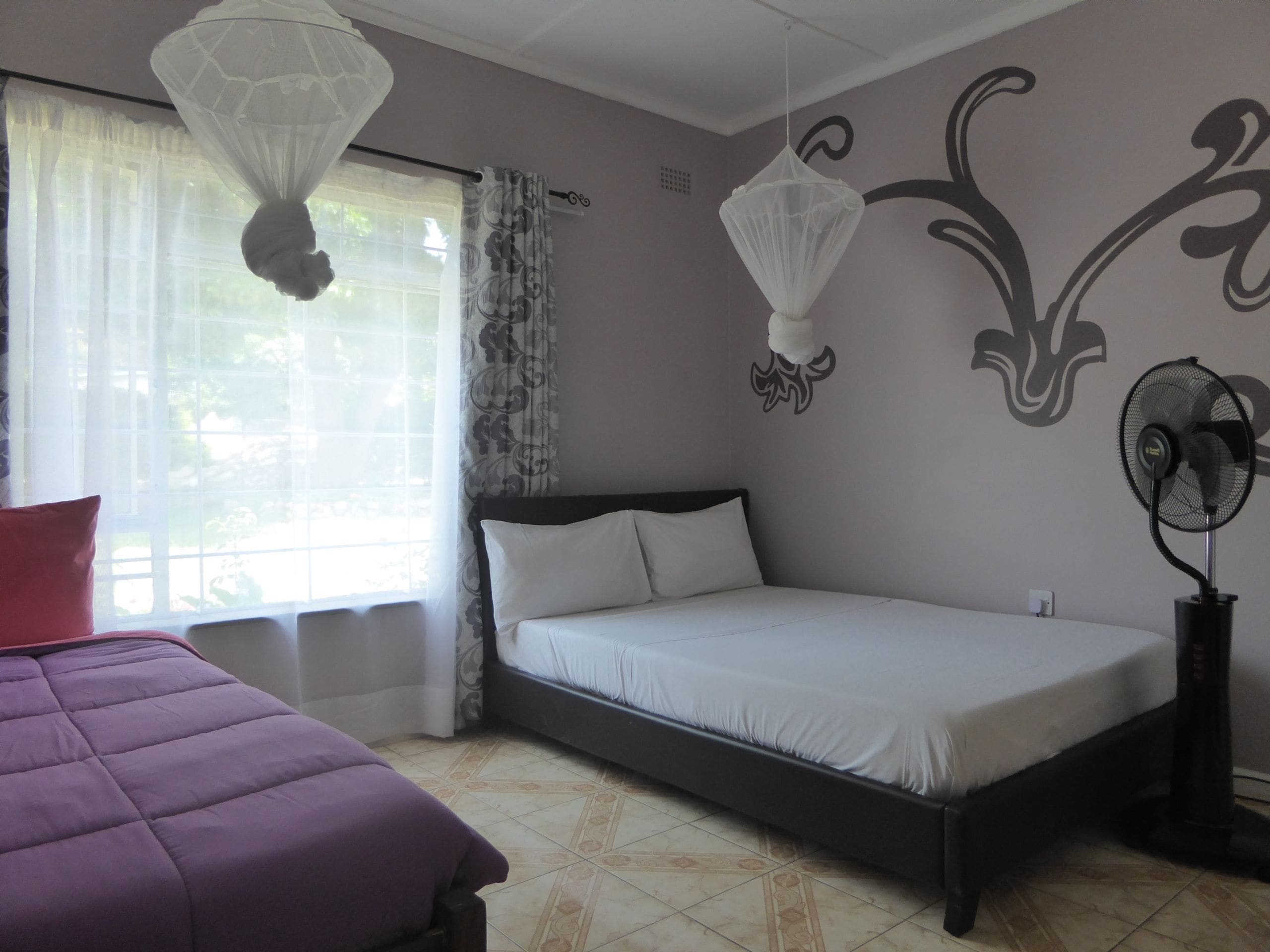 A bedroom with a bed, a bedside table, a window with curtains, a wall-mounted fan, and a decorative wall design.